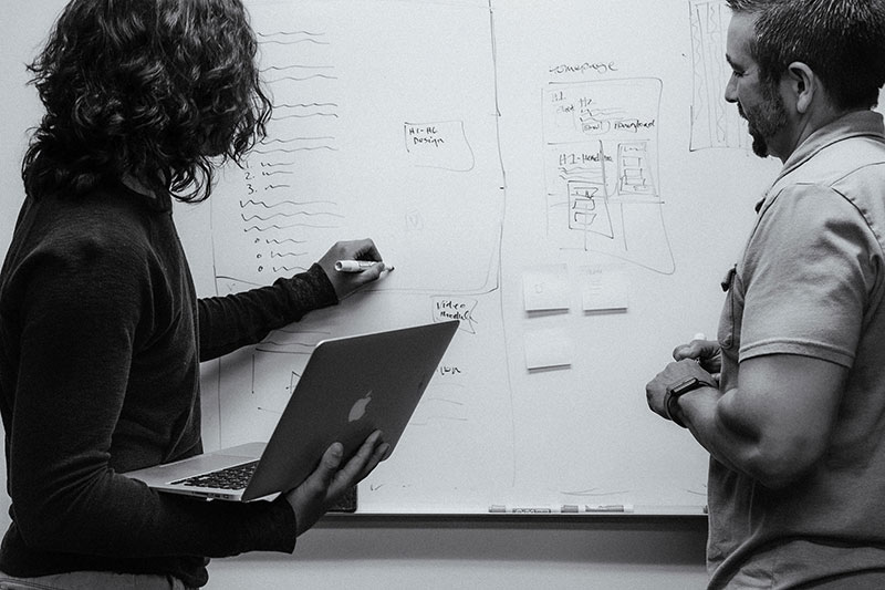 Two people collaborating at a whiteboard; one is writing with a marker while holding a laptop, and the other is observing and engaging in the discussion. The whiteboard is filled with diagrams, lists, and notes, indicating a planning or brainstorming session.