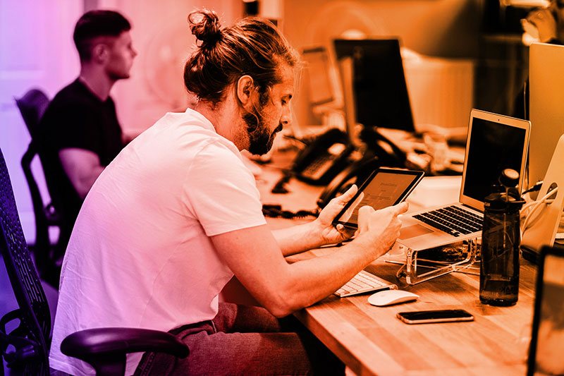 A man with a beard and tied-back hair sits at a desk in an office, working with a tablet while a laptop and other devices are in front of him. Another person works at a computer in the background. The workspace appears modern and technology-focused.