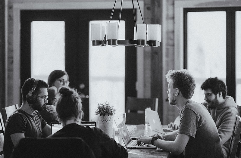 A group of six people work together at a large wooden table in a well-lit room. Some are wearing headphones, and most are focused on laptops. The setting feels collaborative and casual, with natural light streaming through the windows.