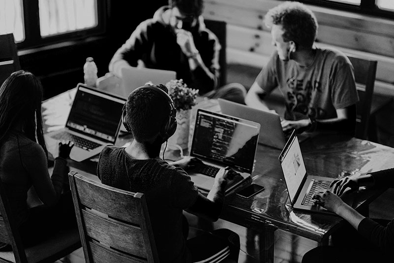 A small team of six people sits around a table with laptops open, deeply focused on their screens. The atmosphere appears tech-driven and productive, with a mix of coding and discussion happening in a shared workspace.