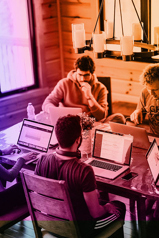 Five individuals are working at a wooden table in a cozy room with natural lighting. Most are on laptops, concentrating or discussing tasks.