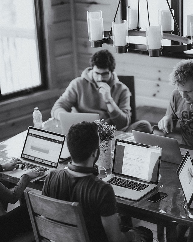 Five individuals are working at a wooden table in a cozy room with natural lighting. Most are on laptops, concentrating or discussing tasks.