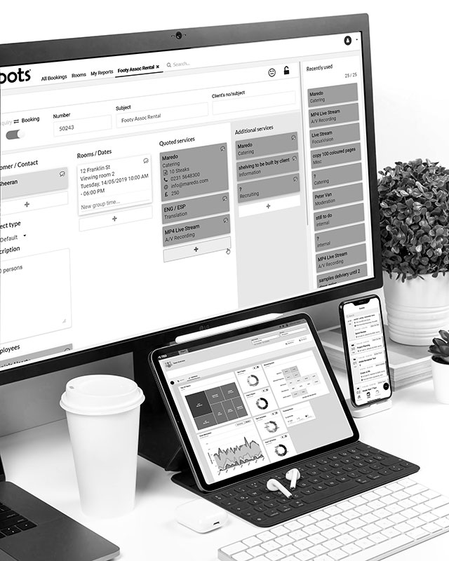 A professional desk setup with a large monitor, tablet, and smartphone, all displaying business dashboard interfaces. The screen shows a booking or scheduling system. The workspace includes a coffee cup, wireless keyboard, earphones, and a potted plant, indicating a digital, organized workspace.